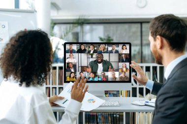 Two business colleagues of different nationalities talking on video call with colleagues. On computer screen is a multi-ethnic group of business partners gathered in an online video conference