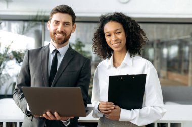 Two successful business people, caucasian man ceo of company, african american female top manager, in formal wear, are standing in modern office, looking at the camera, smiling, partnership concept