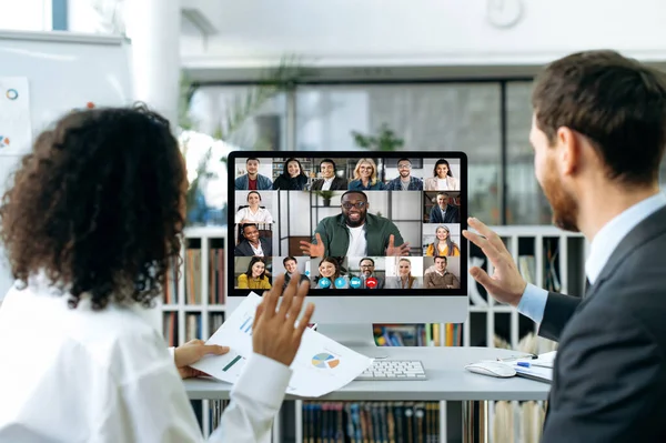 Two business colleagues of different nationalities talking on video call with colleagues. On computer screen is a multi-ethnic group of business partners gathered in an online video conference — Fotografia de Stock