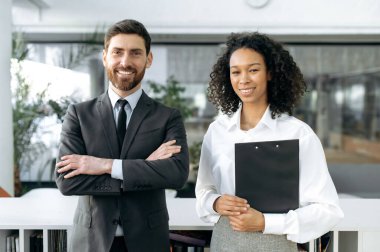 Friendly multiracial business colleagues, African American businesswoman, Caucasian businessman, company workers, standing in a modern office, looking at the camera, smiling