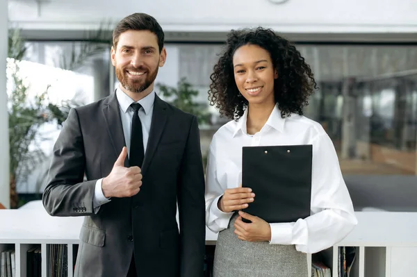 Positive successful multiracial business partners, African American business woman, Caucasian business man, company workers, standing in a modern office, looking at the camera, smiling friendly