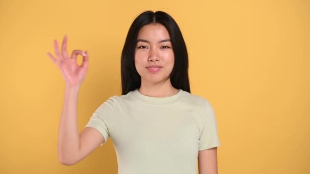 Chinese positive happy young brunette woman, shows to the camera OKAY gesture, standing on isolated orange background, in t-shirt, smiling — Vídeo de Stock