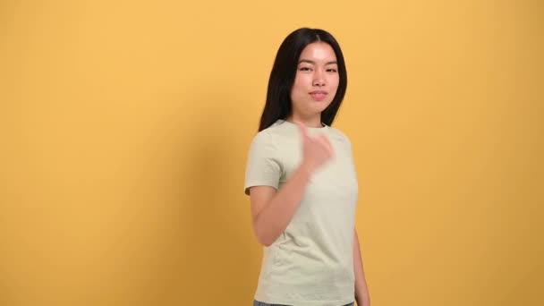 Happy lovely millennial brunette long-haired Asian young woman, points with hands to empty space to back, looks at camera, smiling friendly, stands on isolated background in casual t-shirt — Vídeo de Stock