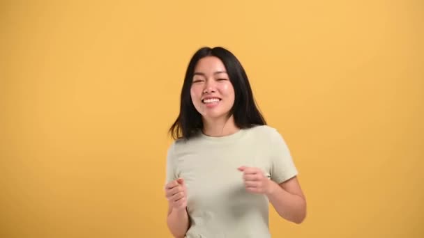 Joy, victory, luck. Cheerful happy chinese brunette girl in casual t-shirt, dancing to favorite music, having fun, rejoice in luck, on isolated orange background — Vídeo de Stock