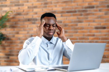 Migraine, fatigue. African American office worker, manager, programmer, tired of working at a laptop in the office, having a headache, massaging temples, closed his eyes, needs rest