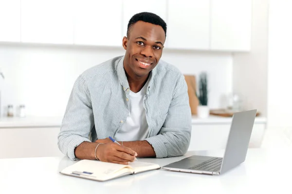 Positive modern black man, freelancer, designer or student, sits at a kitchen at home, using laptop, studying or working online, improves qualifications, smil friendly, takes notes. E-learning concept — 스톡 사진