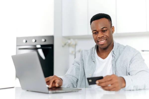 Hombre negro alegre se sienta en la cocina en casa, vestido con ropa casual, utilizando el ordenador portátil y la tarjeta de crédito para las compras en línea, está introduciendo datos para el pago, transacción en línea, orden de entrega a domicilio, sonrisas — Foto de Stock