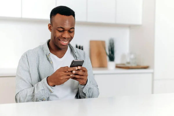즐거운 흑인 남자가 평상복을 입고 부엌에 앉아 인터넷 과 소셜 네트워크를 둘러보고 친구나 가족 과 문자 메시지를 보내고 좋은 소식을 발견 했다. — 스톡 사진