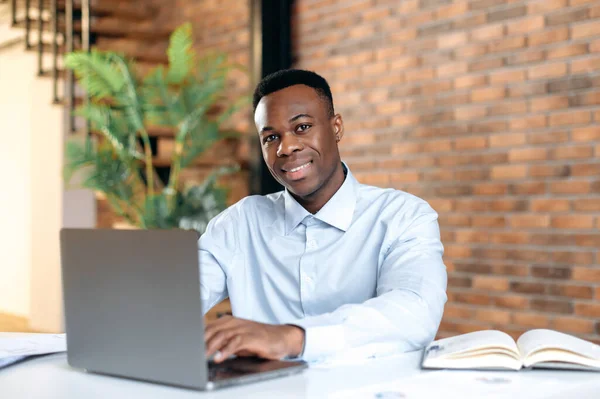 Positive confident black man using laptop in modern office. Portrait successful businessman, freelancer, manager, in formal shirt, working and typing on laptop, looking at camera, smiling friendly — Fotografia de Stock