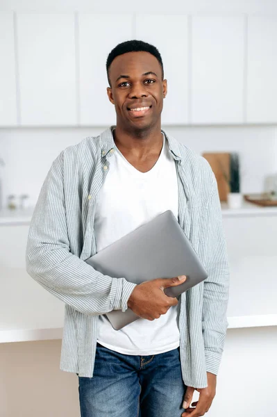 Portrait of African American handsome guy holding laptop in hand, black male student or freelancer, in stylish casual clothes, standing at home in the kitchen, looking at the camera, smiles friendly — 스톡 사진