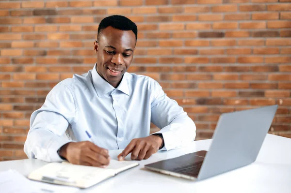 El hombre de negocios afroamericano enfocado que escribe notas en el cuaderno durante el webinar en línea, está mejorando calificaciones. Estudiante negro inteligente estudio en línea, escuchar conferencia en línea, utilizando el ordenador portátil — Foto de Stock