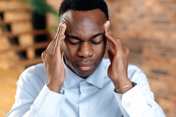 一位精疲力竭的非洲裔美国人在工作时头痛、偏头痛、工作过度，他的特写镜头，悲伤的人生病了，需要治疗和休息，闭上眼睛，按摩太阳穴 — 图库照片
