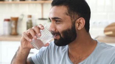 Sağlıklı bir yaşam tarzı. Pozitif sağlıklı, çekici Hintli sakallı adam, mutfakta duruyor, bardaktan temiz su içiyor, yan tarafa bakıyor, sağlığına dikkat ediyor, günlük bir bardak su içiyor, gülümsüyor.