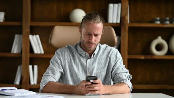 Focused caucasian guy is sitting at a table in a modern office, using his smartphone, chatting with friends on social networks, texting with colleagues, answering email, tired, looking away, need rest — Stockvideo