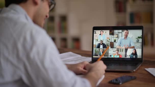 Video call, online conference using app and laptop. Over shoulder view of student or freelancer, studies or works remotely, watches an online lecture, takes notes, multiracial people on laptop screen — Wideo stockowe