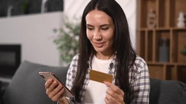 Caucasian young adult beautiful happy brunette woman using mobile phone and bank card to pay for purchases and delivery on Internet, entering credit card details, looking at phone, smiling