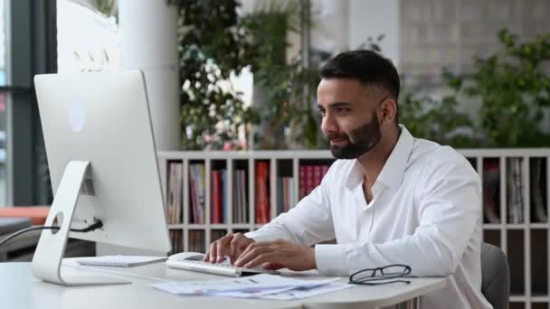 Feliz joven adulto exitoso líder empresarial indio, gerente o trabajador de oficina con barba sentado en la oficina, utilizando la computadora, regocijándose en el éxito o el beneficio, gesticulando con los puños, sonriendo — Vídeo de stock