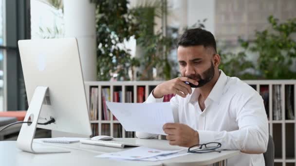 Занят успешный молодой взрослый индийский бизнес-лидер, менеджер или офисный работник с бородой, сидя в офисе, используя компьютер, изучая документы и диаграммы, проводя аналитику, делая заметки — стоковое видео