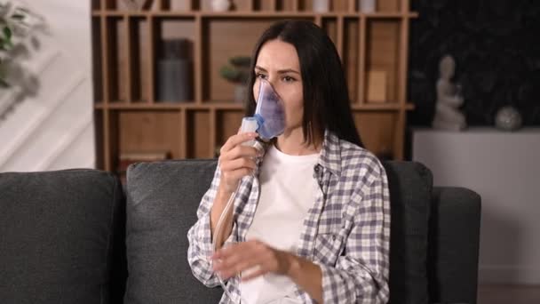 Ill caucasian woman with an inhaler, sick girl doing inhalation at home, she use nebulizer and inhaler for the treatment sitting on the couch at home at quarantine alone — Stockvideo