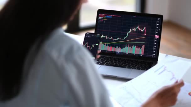 Successful smart woman investor and crypto trader, uses laptop and smartphone, analyzes charts of trading in stock market and digital cryptocurrency exchange, conducts analysis, trading crypto coins — Vídeo de stock