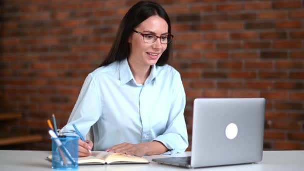 Онлайн деловая встреча. Дружеская позитивная успешная кавказская деловая женщина, топ-менеджер, одетый в рубашку, общается с коллегами или клиентами через видеосвязь, делает заметки, улыбается — стоковое видео