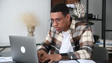 Upset frustrated hispanic guy, office worker or freelancer, sits at work desk, looks sadly at his laptop, failed the deal, got a bad result of work, have trouble, negative mood, worried, confused