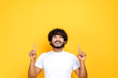Neşeli kıvırcık saçlı, bin yıllık Hintli ya da Arap adam, boş uzayı işaret ediyor, yukarı bakıyor, dostça gülümsüyor, izole edilmiş turuncu arka planda duruyor, günlük beyaz tişörtle, sahte konsept.