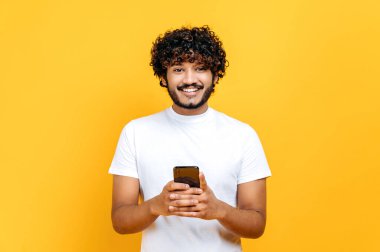 Beyaz tişörtlü, heyecanlı, yakışıklı Hintli adam, akıllı telefon kullanıyor, internetten arkadaşlarıyla mesajlaşıyor, sosyal ağlara, haberlere bakıyor, kameraya bakıyor, gülümsüyor, izole edilmiş turuncu arka planda duruyor.