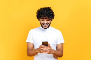 Heyecanlı kıvırcık saçlı, sıradan beyaz tişörtlü, akıllı telefonunu kullanan, arkadaşlarıyla internetten mesajlaşan sosyal ağlara, haberlere, gülümseyen, izole edilmiş turuncu arka planda duran,