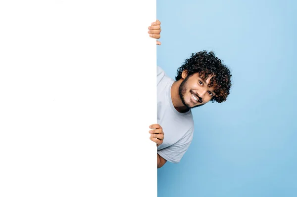 Positive Indian or Arabian guy, in casual t-shirt, peeking out from behind advertisement whiteboard, demonstrating blank copy space for your text or design, isolated blue background, mock-up concept — Stock Photo, Image