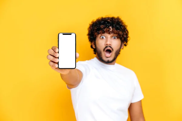 Rozostřený překvapený indický mladý muž v bílém tričku, drží smartphone s maketou prázdné bílé obrazovky, stojí na izolovaném oranžovém pozadí, šokované pohledy do kamery — Stock fotografie