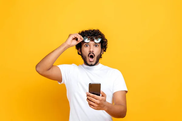 Překvapený šokovaný indián v bílém tričku, drží v ruce smartphone, sundá si brýle a s úžasem se podívá do kamery, obdržel nečekanou zprávu nebo zprávy, izolované oranžové pozadí — Stock fotografie