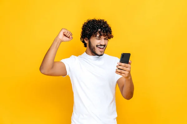 Úžasný radostně vzrušený indický nebo arabský chlap drží smartphone, získat nečekané zprávy, výhra loterie, stojí na izolovaném oranžovém pozadí, veselý výraz obličeje, zubatý úsměv, gestikulace s pěstí — Stock fotografie