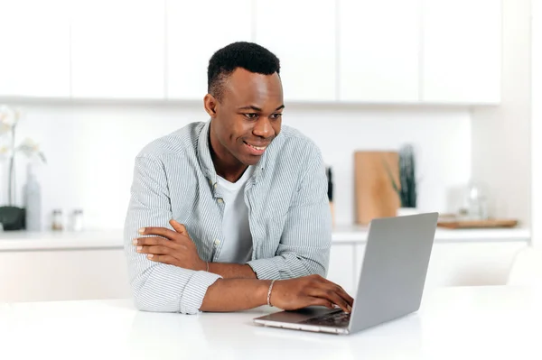 Agradable hombre negro moderno, freelancer, diseñador o estudiante, sentado en casa en la cocina, utilizando el ordenador portátil para estudiar o trabajar en Internet mejorando sus habilidades, sonriendo amigable. Concepto de aprendizaje electrónico — Foto de Stock