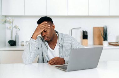 Stresli, esmer tenli, serbest çalışan, mutfakta bir masada oturan, yorgunluk, baş ağrısı, migren, başını tutup gözlerini kapatan, dinlenmeye ihtiyacı olan, bilgisayar başında çok çalışan bir adam.