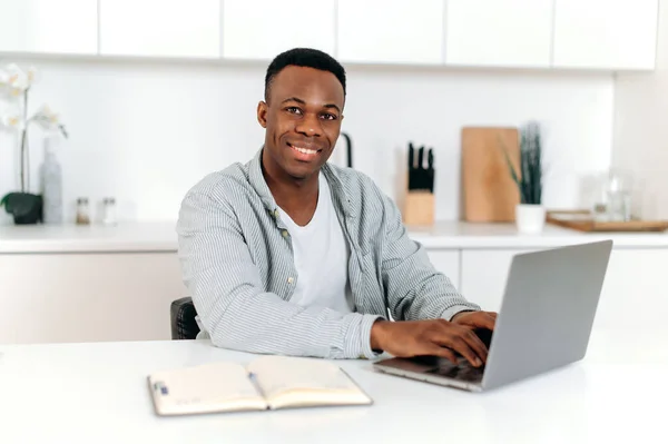 친절 한 흑인, 프리랜서, 디자이너 또는 학생, 부엌에 앉아 노트북 과 수첩을 사용하거나 인터넷 서핑을 하고, 기술을 향상시키고, 다정하게 웃는다. E - 학습 개념 — 스톡 사진