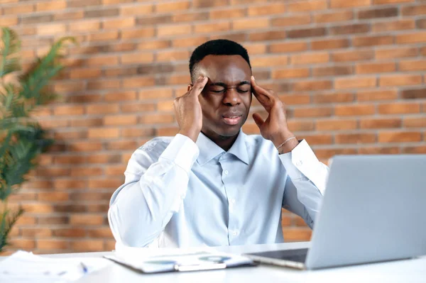 Overworked exhausted young adult African American man, manager or financial leader, stressed a d tired from computer work in the office, massages his temples, closes his eyes, needs rest, treatment — Stock Photo, Image