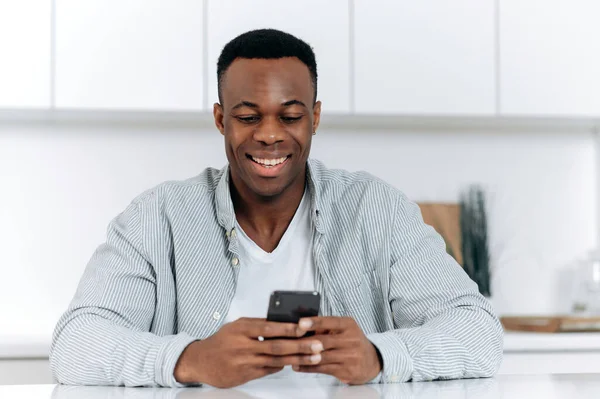 아프리카 계 미국인 매력적 인 긍정적 인 청년은 평상복을 입고 부엌에서 식탁에 앉아 여가 시간을 보내며 스마트폰을 사용하고 인터넷을 둘러보고 소셜 미디어를 통해 수다를 떨며 미소를 짓는다. — 스톡 사진