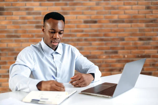 Un joven afroamericano mantiene una reunión de negocios con sus colegas. Director o gerente de la compañía inteligente de éxito lindo usando el ordenador portátil y el bloc de notas para la negociación encore con colegas o clientes. — Foto de Stock