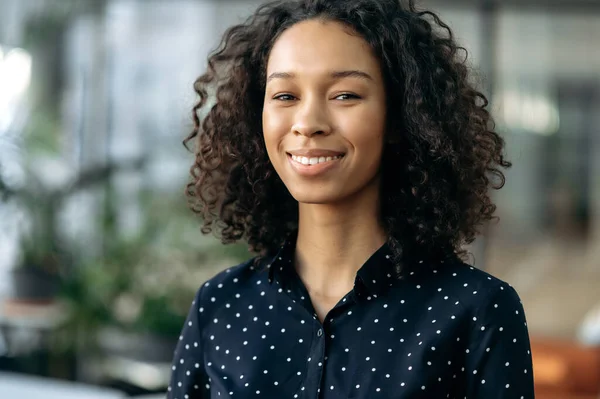 Close-up of curly atraktif positif african american girl, nice young business woman, student or freelancer, real estate agent, standing in office, looking at the camera, smiling friendly — Stok Foto