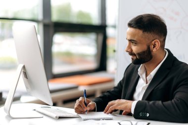 Zeki, başarılı Hintli takım elbiseli başarılı iş adamı, şirket müdürü, finansal danışman veya komisyoncu masasında oturuyor, dizüstü bilgisayarda çalışıyor, çevrimiçi konferans dinliyor, notlar alıyor, ekrana bakıyor.