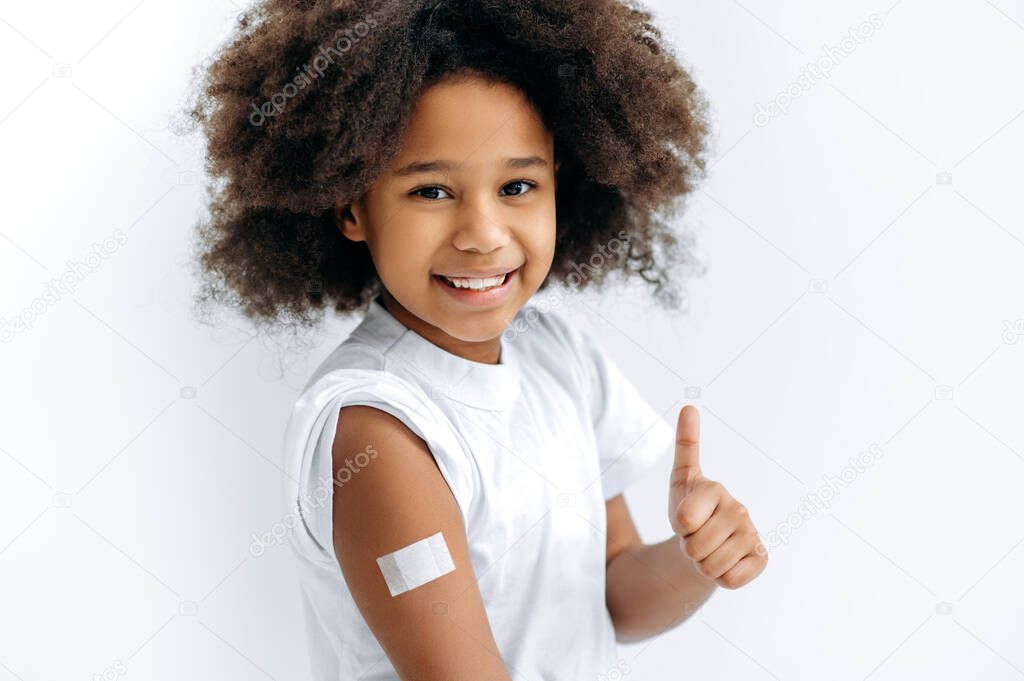 Happy African American cute little girl, preschooler, with a band-aid on her shoulder after vaccination, for immunity against covid 19, and other diseases, stand on isolated white background, thumb-up