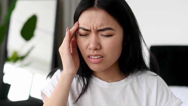 Droevig gefrustreerde aziatische brunette jonge vrouw in stress, zitten op de bank alleen thuis, ervaren hoofdpijn, migraine, masseren haar slapen, sluiten van haar ogen, behoefte aan rust en behandeling — Stockvideo