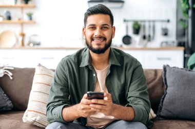 Mutlu Hintli ya da Arap bir adam, rahat giyiniyor, evinde oturuyor, akıllı telefonunu kullanıyor, mesajlaşıyor, sosyal ağlara takılıyor, arkadaşlarıyla mesajlaşıyor, kameraya bakıyor, arkadaşça gülümsüyor.