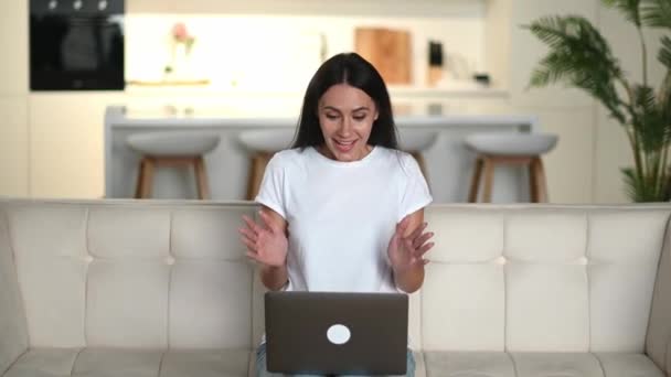 Joyeux jeune femme caucasienne brune confiante et mignonne, en vêtements décontractés, assise sur un canapé, utilisant un ordinateur portable, célébrant un bon profit, réjouissant les nouvelles ou le message, geste avec les poings, regardant l'écran, sourire — Video