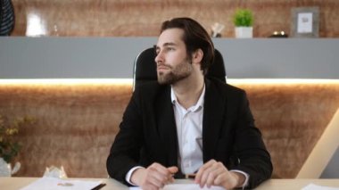 Başarılı, zeki, Kafkasyalı bir iş adamının video portresi, şirket CEO 'su, resmi bir takım elbise giyiyor, kameraya dostça bakıyor, modern ofisteki işyerinde otururken.