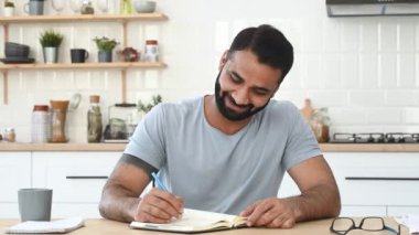 Zeki, pozitif Hintli bir adam, evdeki mutfakta bir masada oturuyor, konferans dinliyor, video konferansı yapıyor, not defterine notlar alıyor. Uzaktan öğrenme, ileri eğitim