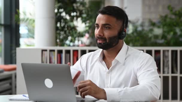 Sociable attrayant entrepreneur intelligent indien, homme d'affaires, directeur des ventes, commerçant, avec écouteurs assis à table dans le bureau, communique par appel vidéo avec des collègues ou des clients, salutation, sourires — Video