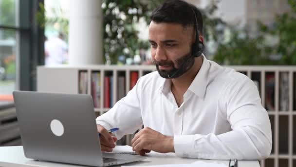 Inteligente propositado atraente indiano homem com fone de ouvido, gerente, trabalhador de suporte, consultor on-line, executivo sentado no laptop no escritório moderno, ouvir, gravar apelo, fazendo consulta on-line — Vídeo de Stock