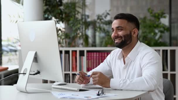 Φιλική αυτοπεποίθηση επιτυχημένος Ινδός, επιχειρηματίας ή μέντορας, κάθεται στο γραφείο του, φορώντας ένα λευκό πουκάμισο, χρησιμοποιώντας υπολογιστή για μακρινή επικοινωνία, μιλώντας με έναν συνάδελφο μέσω βιντεοκλήσης, χαμογελώντας — Αρχείο Βίντεο